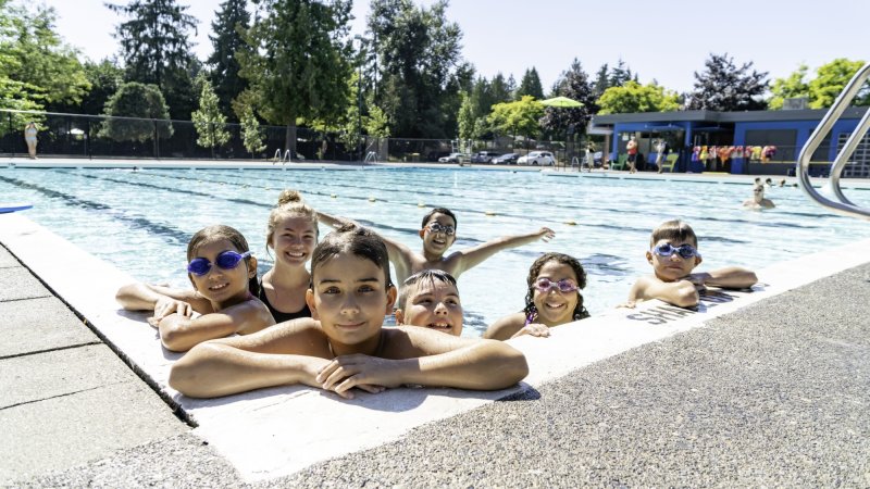 Group of children with swim goggles, smiling and enjoying themselves at an outdoor pool on a sunny day.