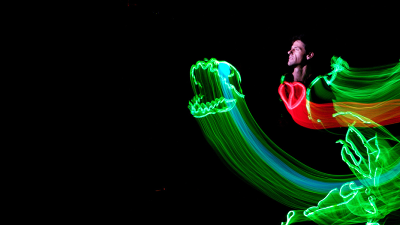 you see a performer's face amongst swirls of green, blue and red coloured lights depicting a dinosaur with the red light as a heart by the performers chest. 