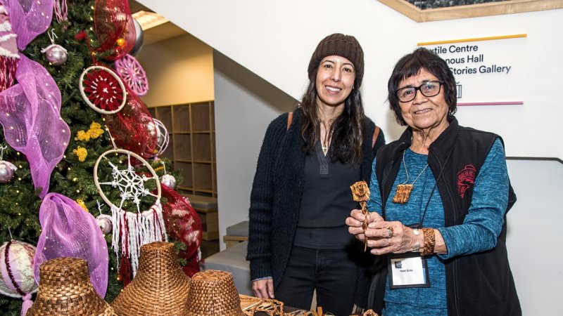  Two women stand smiling behind a display table at a craft market, with a Christmas tree in the background. 