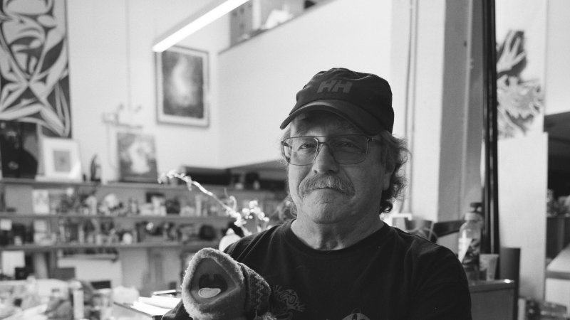 A black and white photograph of a man holding a frog puppet in his arms. He wears glasses and a baseball cap. He has a smile and a moustache. He wears a tee shirt with frogs. He is in a workroom.