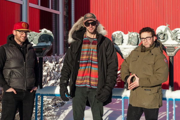 Image of three band members outside in front of a red wall wearing winter clothing 