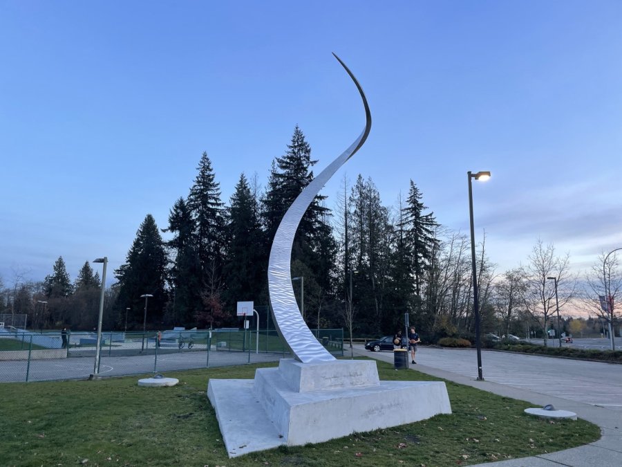 A spiraling metallic sculpture stands in an evening park setting, with trees, a tennis court, and a passerby in view.