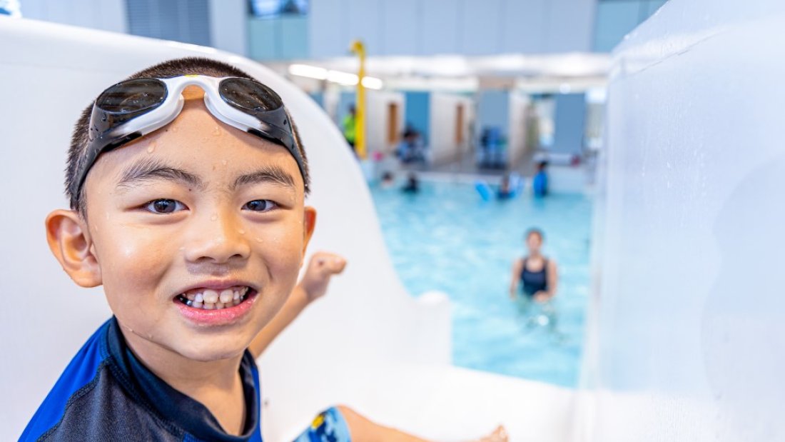 A boy smiling with goggles at the top of a slide in a pool.