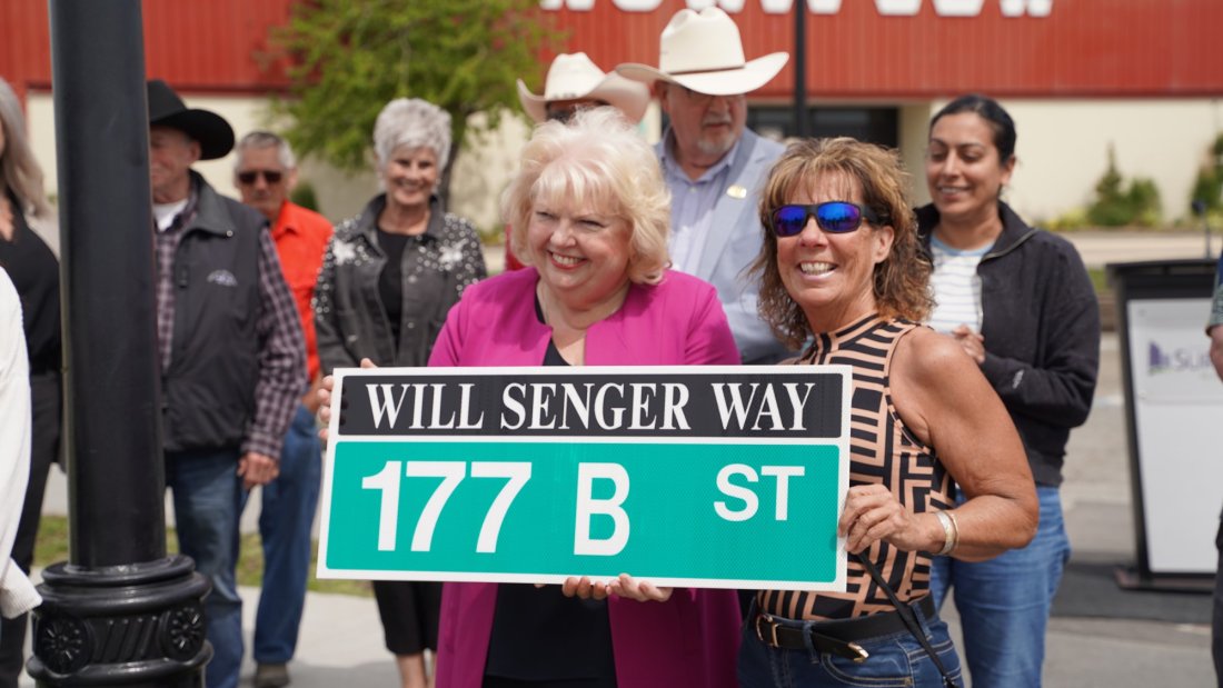 Two women smiling and holding a new street sign "Will Senger Way" during a street renaming event in front of the Agriplex building.