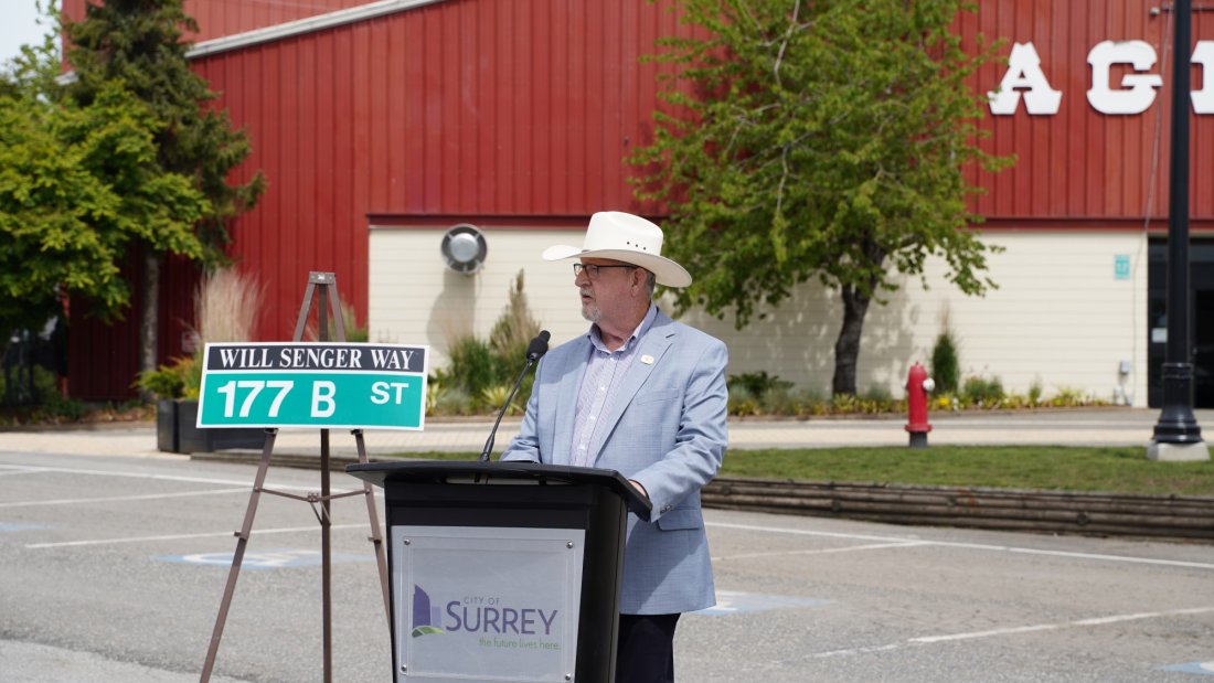 A man in a cowboy hat giving a speech at a podium during a street renaming ceremony, with the new "Will Senger Way" street sign displayed in front of the Agriplex building.