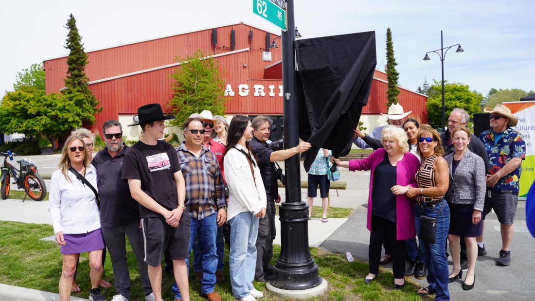 A group of people gathered around a street pole, smiling and preparing to unveil a new street sign, with the Agriplex building in the background.