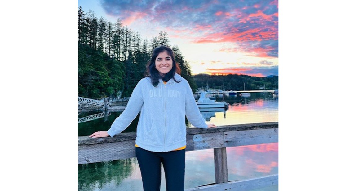 Photo of Kalyani Phadke standing against a wooden bridge, smiling at the camera. A beautiful sunset is seen reflected on the water.