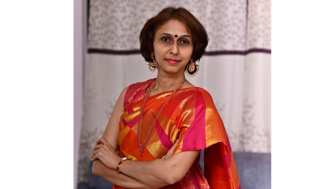 Artist Priya Seetharaman faces the camera with a slight smile on her face. She is wearing a bright red, orange, and pink sari with jewellery.