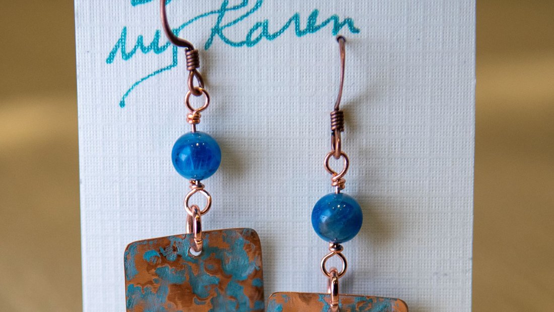 A pair of earrings on a paper backing with a small blue circular bead and a blue and copper rectangle below.