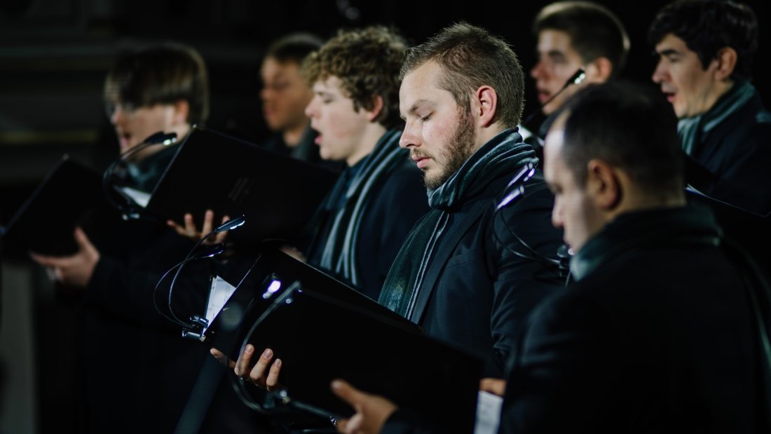 Male choir members in black attire with scarves holding sheet music, focused on singing.