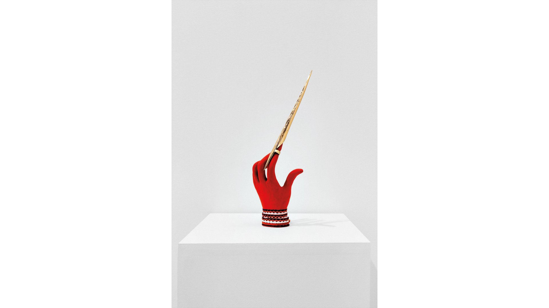 A clay hand painted red stands on a white plinth in a white room wearing several bracelets. A long pointed stick is attached to the index finger.