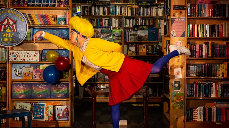 the librarian wearing a yellow cardigan, red skirt and blue tights is in the library in front of bookshelves with lots of books, is balancing on one leg with the other let outstretched and arm in the air