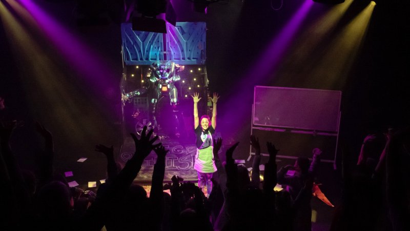 performer on stage with arms up in the air, theatre is dark but for purple lighting, in foreground the audience have their hands up in the air too