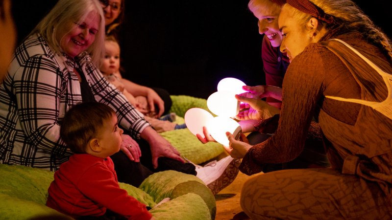 audience of adults and babies sitting on pillows as the performers hold illuminated eggs in front of a baby 