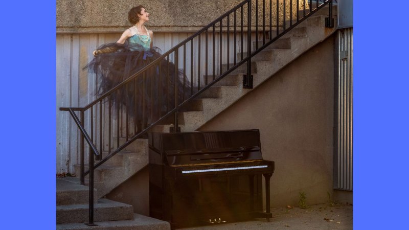 Sarah Hagen stands on a black iron stairwell gazing upwards wearing a large tulle tutu, while a piano is at the base of the stairs.