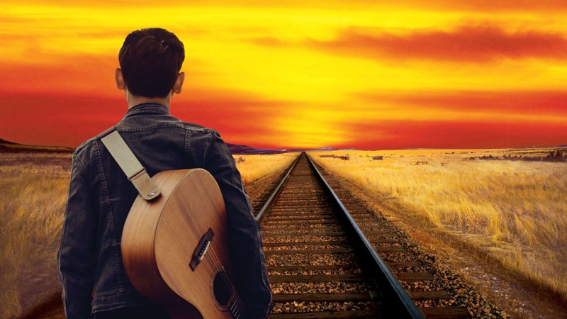 A man with a guitar over his shoulder stands on a train track and looks into the sunset