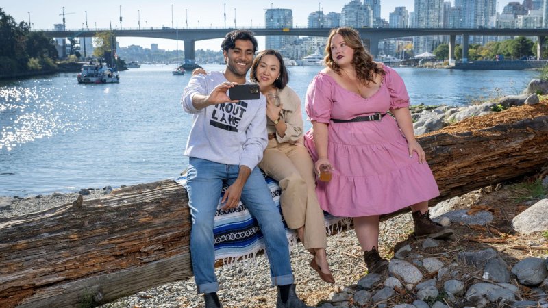 The cast of Someone Like You sit on a log on the beach with the sea behind them talking a selfie.