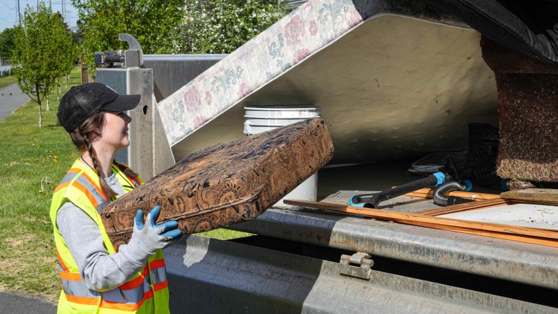 A female individual in a reflective vest and gloves loading an old ornate cushion into a disposal bin during a community cleanup event.