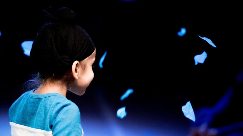 dark background with blue pieces of paper falling around a young child who wears a blue shirt 