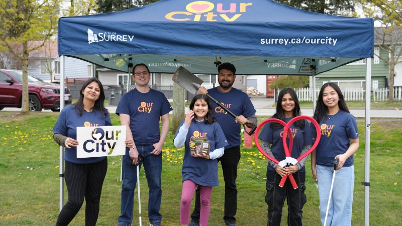  A group of volunteers representing the "Our City" initiative in Surrey, dressed in themed T-shirts, under a community event tent.