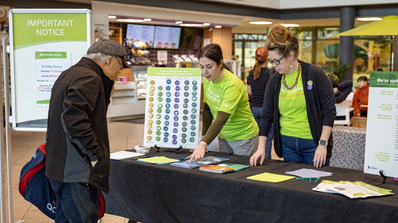 This photo captures a public engagement event with booth attendees in green shirts providing information to a visitor, set against informational displays about a community planning project.
