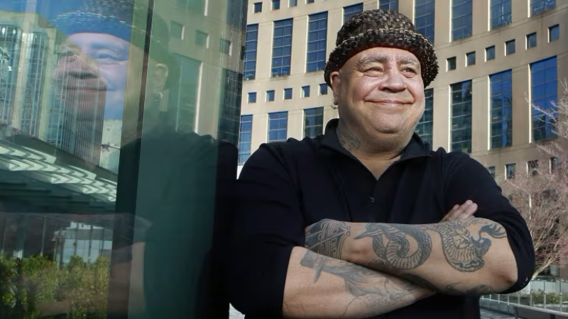 Joseph Dandurand is wearing a hat and stands outside with the vancouver library in the background, his tattooed arms folded across his chest 