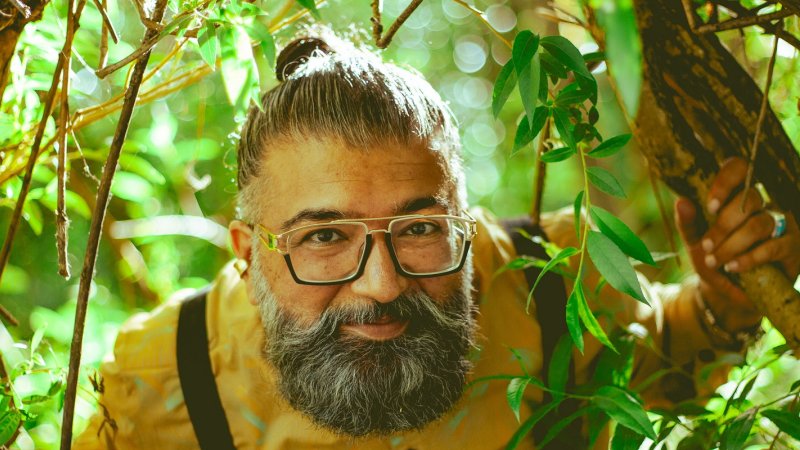 Performer Ruby Singh is a South Asian man who has a beard and wears glasses, he's looking at the camera, surrounded by branches and leaves