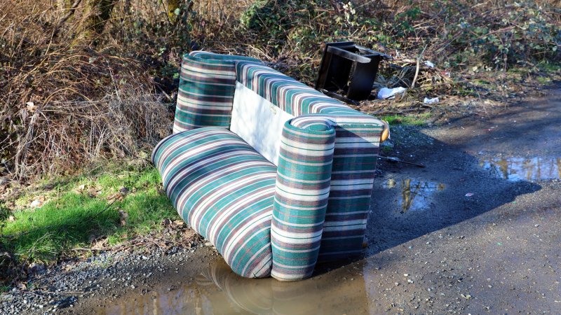 Discarded striped couch and a black trash bin left by the side of a muddy road in a wooded area, highlighting an instance of illegal dumping.