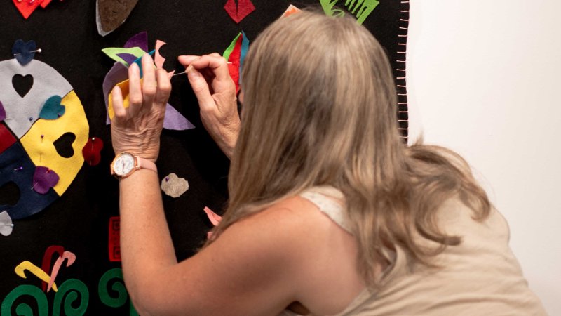 A crouching woman attaches a piece of felt to a blanket hanging on the wall.