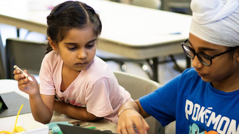 A girl and a boy sit beside each other at a table doing art with paper and crayons.