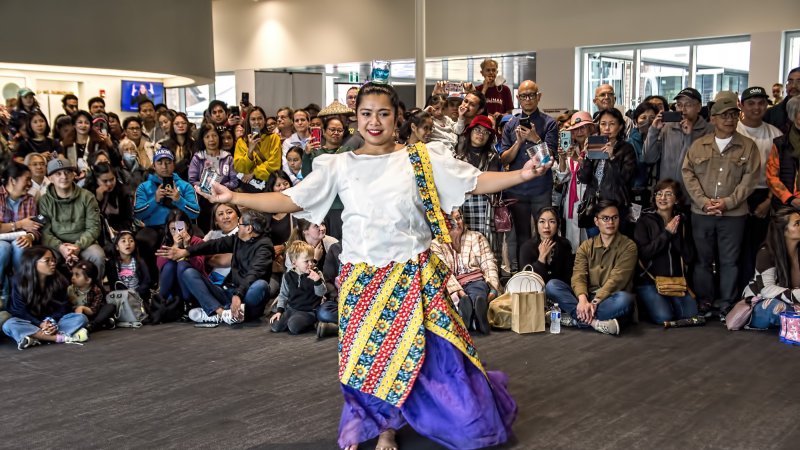 Filipino female dancer in traditional attire performing at an indoor community event with a large, diverse audience watching and taking photos.