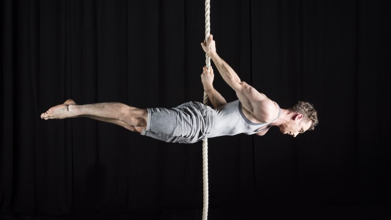 A male acrobat performs a horizontal pose on a rope, with a black background.