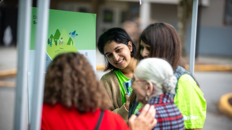 A community engagement event with a smiling facilitator discussing information with attendees near a display board.