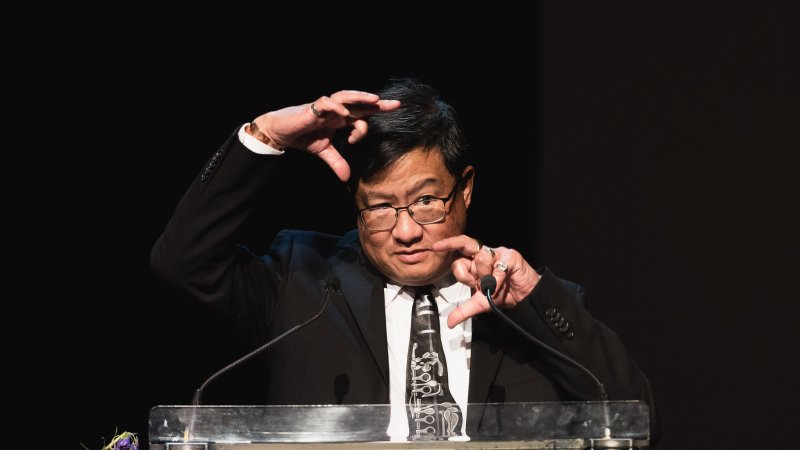 Walter Quan stands behind a podium in a suit and tie making a face and doing expressive hand gestures