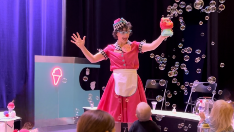 Actor dressed in a 1950s diner style waitress costume stands on the stage in front of audience while she holds a bubble making machine with bubbles coming out of it.