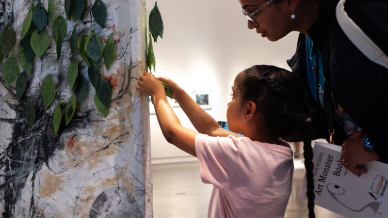 A child pins a leaf onto a canvas with a textured tree image in a gallery space while a caregiver stands behind, leaning in to watch.