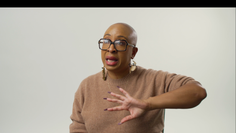 Negarra A. Kudumu wears a beige sweater, dark rimmed glasses, and chunky gold earrings. She is sitting down facing the camera against a neutral wall, and her hand is gesturing in front of her.