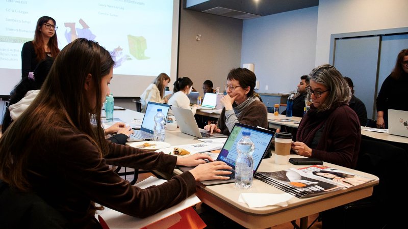 A group of people work on laptops around rectangular beige tables in a room. A person stands at the front in front of a projected image on a screen.