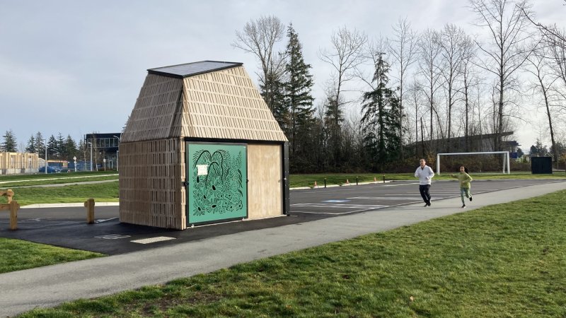 A small wooden structure with artistic designs, situated in a park with people jogging nearby.