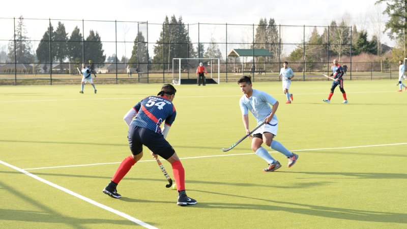 Field hockey players in action during a game on an outdoor turf field, with a goalie and other players in the background.