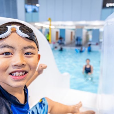 A boy smiling with goggles at the top of a slide in a pool.