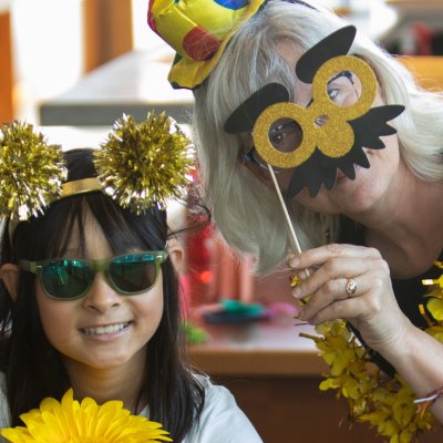 a child wearing sunglasses and a glittery gold pom pom headband and a woman holding up a photo prop of gold glasses and a moustache pose for the camera