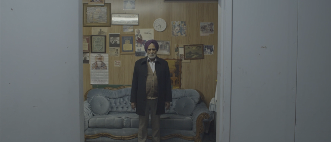  A still from a digital video shows an elderly person standing, facing the viewer. They wear a purple dastar, a coat, a vest, and a white button-up underneath. They are standing inside of a room with an open door. Inside the room is a wood panelled wall with various frames and paper posters. A round clock tells the time. Behind is a velvet-blue couch. This person is not wearing shoes, just socks. They stand on carpeted floor.