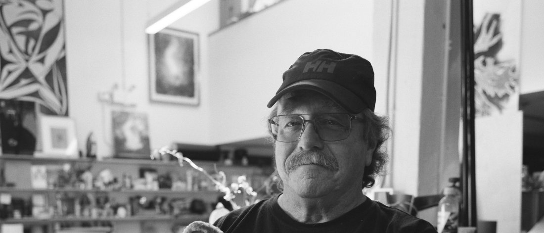 A black and white photograph of a man holding a frog puppet in his arms. He wears glasses and a baseball cap. He has a smile and a moustache. He wears a tee shirt with frogs. He is in a workroom.