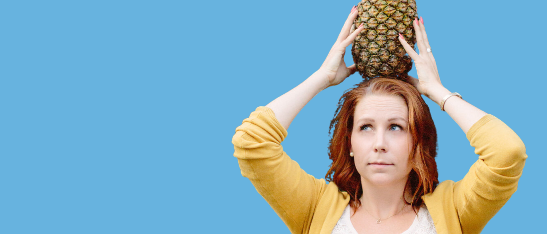 Katie-Ellen is wearing a yellow cardigan and holding a large pineapple on her head