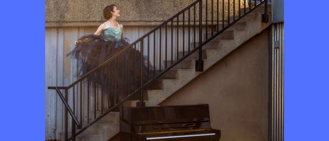 Sarah Hagen stands on a black iron stairwell gazing upwards wearing a large tulle tutu, while a piano is at the base of the stairs.