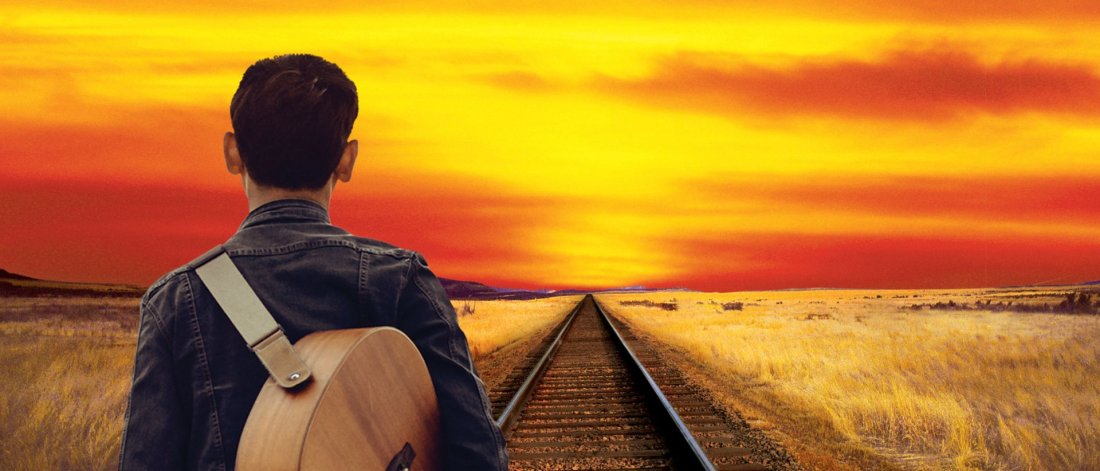A man stands on a railroad track looking at the sunset with a guitar strapped to his back.