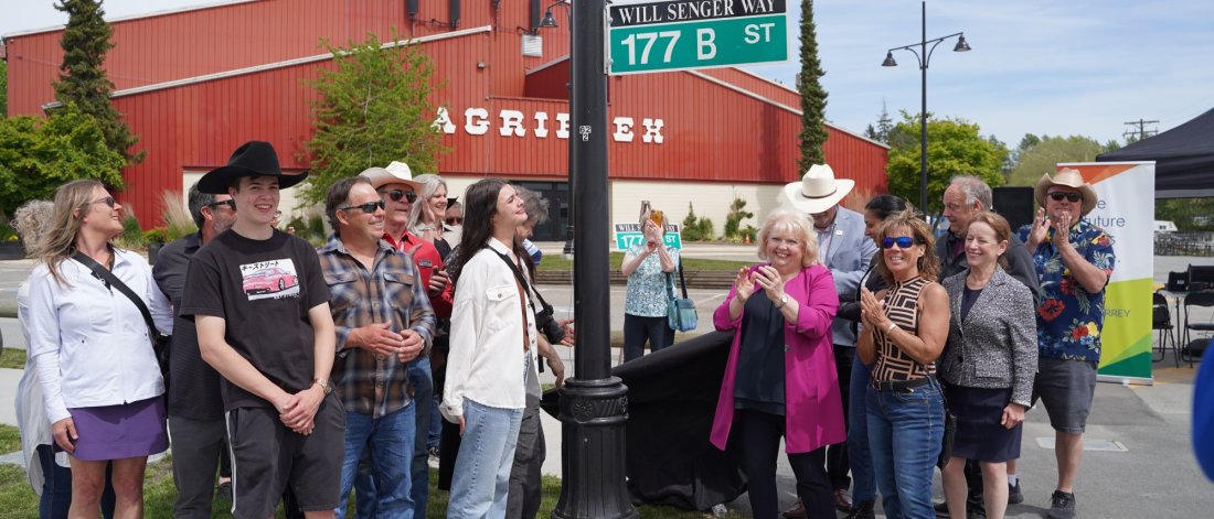 A group of people clapping and celebrating at a street renaming ceremony, with a "Will Senger Way" street sign unveiled, in front of the "Agriplex" building.