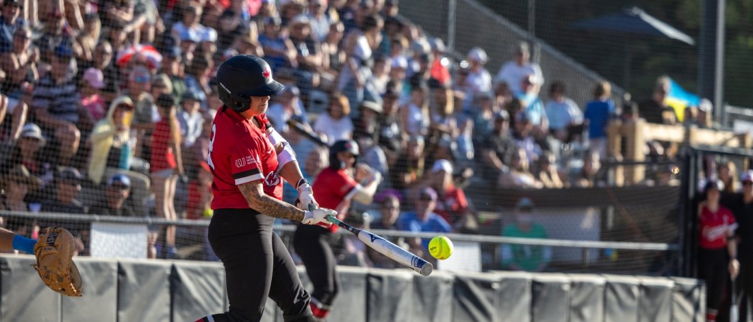 A Team Canada softball player in action, swinging at a pitch during a game, with spectators in the background.