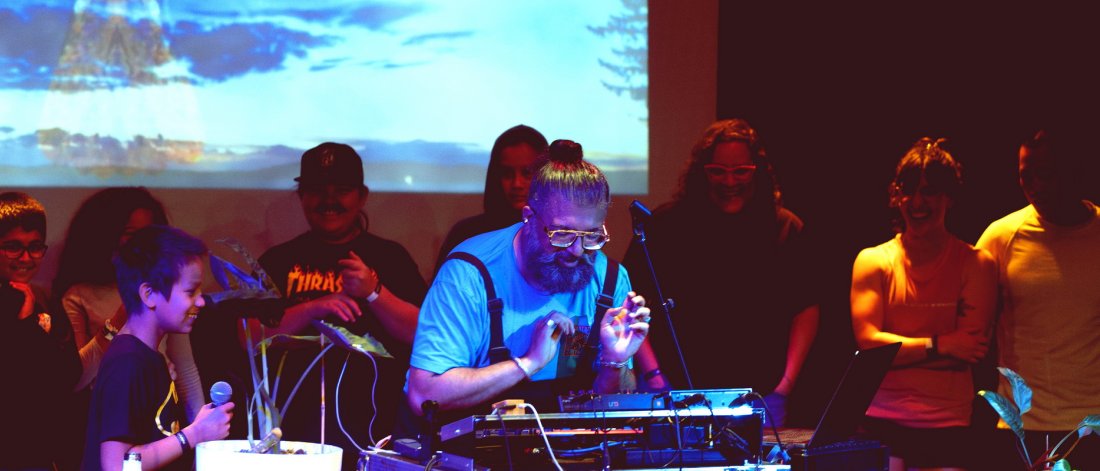 Ruby Singh (aka Ruploops) on performing on stage, projection in the background, audience of all ages around him, young person holding a microphone